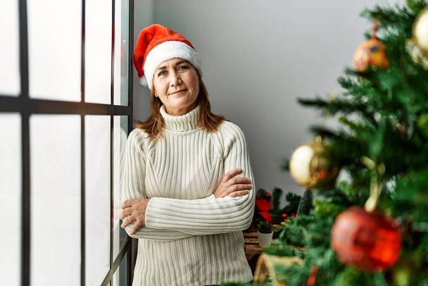 Kaukasische Frau mittleren Alters lächelt selbstbewusst und steht mit verschränkten Armen am heimischen Weihnachtsbaum - Foto, Bild