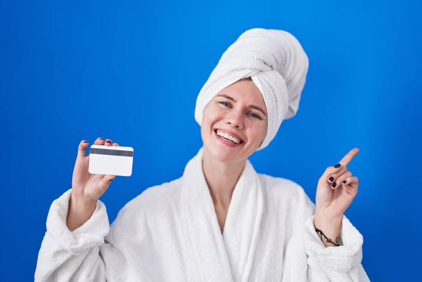 Mulher branca loira vestindo roupão segurando cartão de crédito sorrindo feliz apontando com a mão e o dedo para o lado  - Foto, Imagem