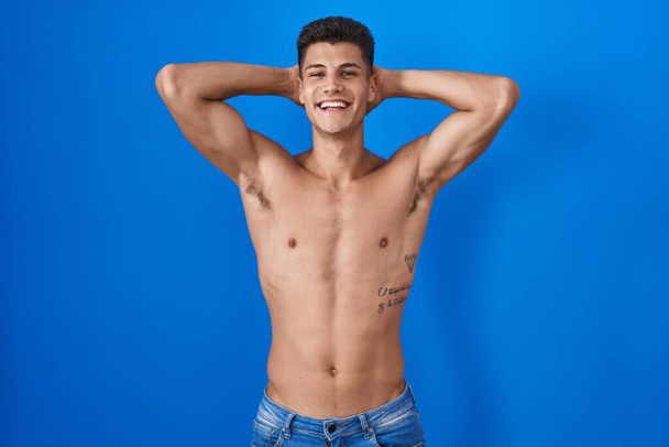 Jeune homme hispanique debout torse nu sur fond bleu relaxant et étirant, bras et mains derrière la tête et le cou souriant heureux  - Photo, image
