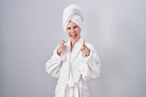 Bornoz giymiş sarışın beyaz kadın mutlu ve komik yüzlü parmaklarını kameraya doğrultuyor. iyi enerji ve titreşimler.  - Fotoğraf, Görsel