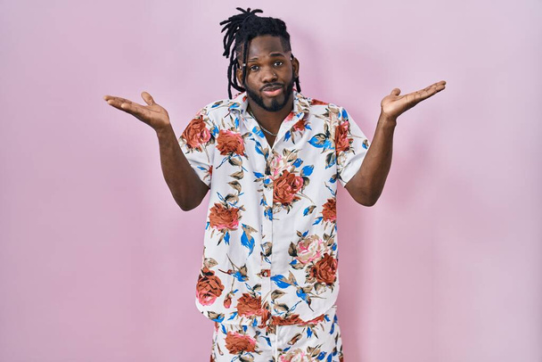 Afrikanischer Mann mit Dreadlocks, Sommerhemd über rosa Hintergrund, ratlos und verwirrt mit erhobenen Armen und Händen. Zweifelhaftes Konzept.  - Foto, Bild