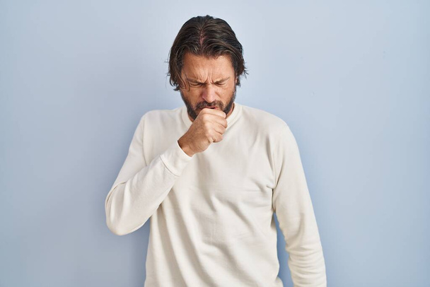 Przystojny mężczyzna w średnim wieku ubrany w luźny sweter na niebieskim tle źle się czuje i kaszle jako objaw przeziębienia lub zapalenia oskrzeli. koncepcja opieki zdrowotnej.  - Zdjęcie, obraz