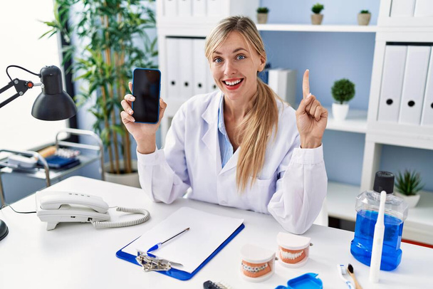Belle dentiste femme montrant écran smartphone souriant avec une idée ou une question pointant du doigt avec le visage heureux, numéro un  - Photo, image