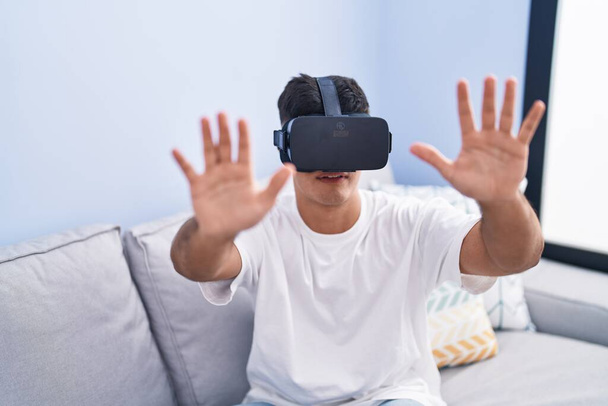 Jeune homme hispanique jouant à un jeu vidéo en utilisant des lunettes de réalité virtuelle à la maison - Photo, image