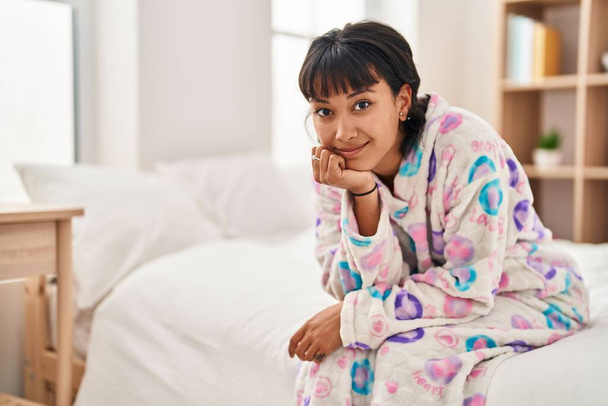 Junge schöne hispanische Frau lächelt zuversichtlich im Schlafzimmer auf dem Bett sitzend - Foto, Bild