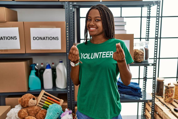 Fiatal afro-amerikai nő, aki önkéntes pólóban dolgozik adománygyűjtő standon, és pénzt gesztikulál a kezével, fizetést kér, milliomos üzletet.  - Fotó, kép