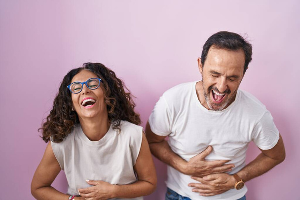 Edad media pareja hispana juntos sobre fondo rosa sonriendo y riendo duro en voz alta porque broma loca divertida con las manos en el cuerpo.  - Foto, imagen
