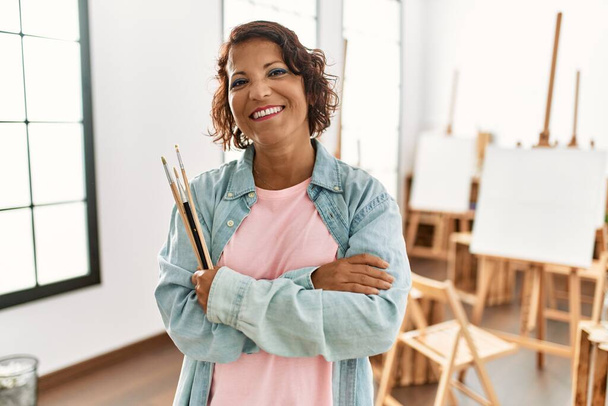 Latynoska artystka w średnim wieku uśmiecha się szczęśliwa stojąc z rękami skrzyżowanymi w pracowni artystycznej. - Zdjęcie, obraz