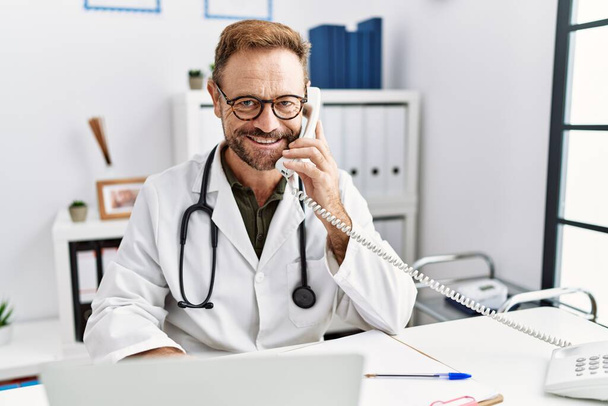 Middle age hispanic man wearing doctor uniform talking on the telephone at clinic - 写真・画像
