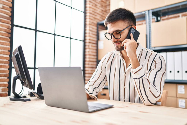 Giovane uomo ispanico e-commerce business worker utilizzando computer portatile parlando su smartphone in ufficio - Foto, immagini