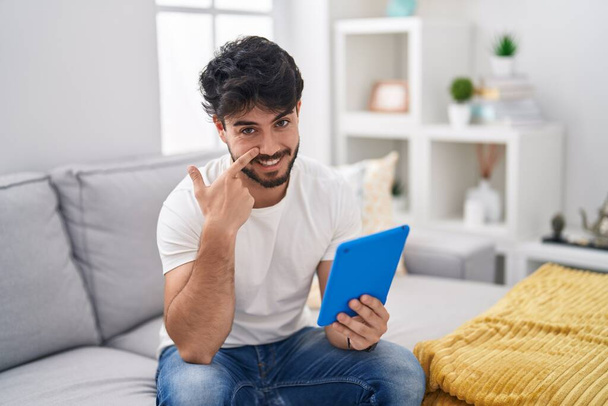 Hispanic man with beard using touchpad sitting on the sofa pointing with hand finger to face and nose, smiling cheerful. beauty concept  - Φωτογραφία, εικόνα