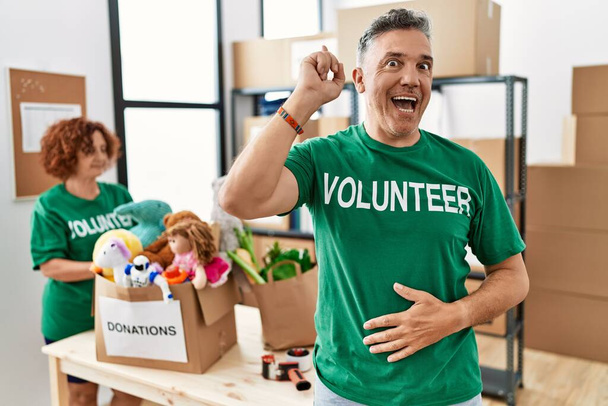 Middle age man wearing volunteer t shirt at donations stand pointing finger up with successful idea. exited and happy. number one.  - Photo, Image