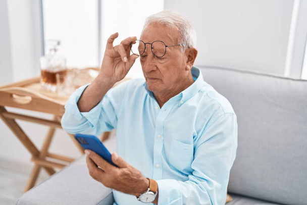 Senior grijs-harige man met behulp van smartphone zitten op de bank thuis - Foto, afbeelding