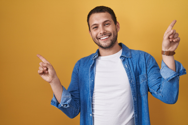 Hispanic man standing over yellow background smiling confident pointing with fingers to different directions. copy space for advertisement  - Valokuva, kuva