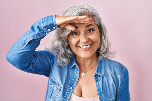 Frau mittleren Alters mit grauen Haaren vor rosa Hintergrund sehr glücklich und lächelnd mit der Hand über dem Kopf in die Ferne blickend. Suchkonzept.  - Foto, Bild