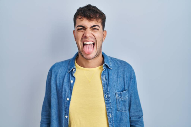 Joven hombre hispano de pie sobre fondo azul sacando la lengua feliz con expresión divertida. concepto de emoción.  - Foto, imagen