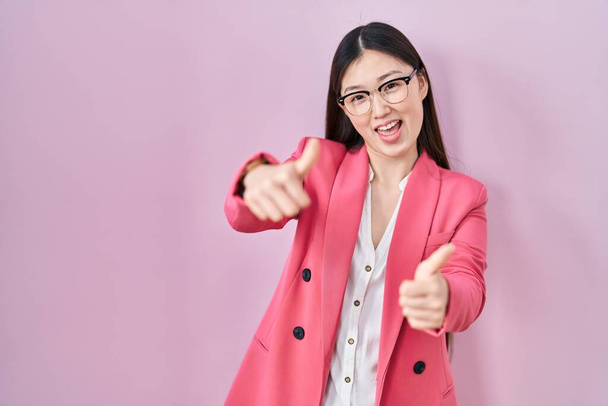 Entreprise chinoise jeune femme portant des lunettes approuvant faire un geste positif avec la main, pouces levés souriant et heureux pour le succès. geste gagnant.  - Photo, image