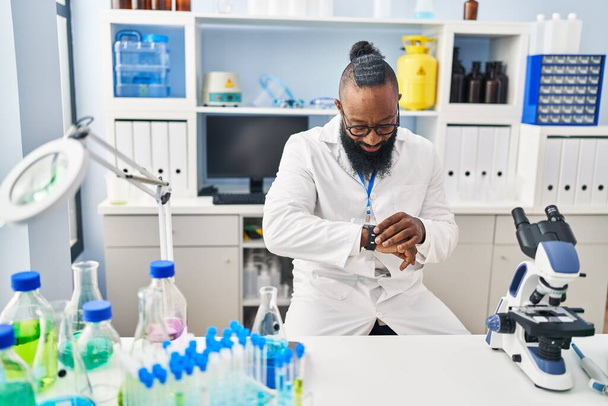 Hombre afroamericano joven vistiendo uniforme científico mirando reloj en el laboratorio - Foto, Imagen