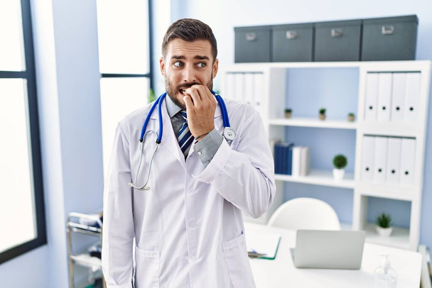 Handsome hispanic man wearing doctor uniform and stethoscope at medical clinic looking stressed and nervous with hands on mouth biting nails. anxiety problem.  - Foto, immagini