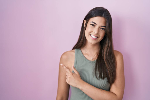 Hispanic woman standing over pink background cheerful with a smile of face pointing with hand and finger up to the side with happy and natural expression on face  - Foto, immagini