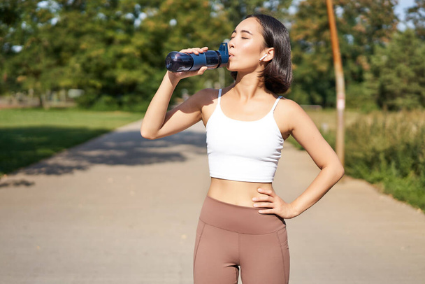 Szczęśliwy azjatycki sportowiec, biegacz pije wodę z butelki podczas biegania, trening na świeżym powietrzu w parku. - Zdjęcie, obraz