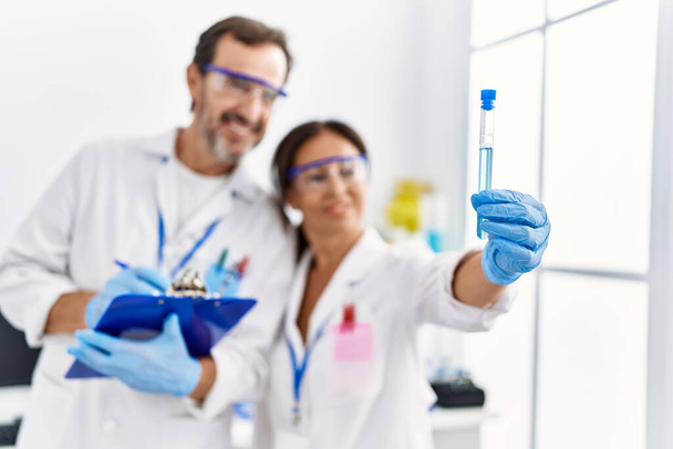 Mężczyzna i kobieta w średnim wieku partnerzy noszący mundur naukowca piszący na schowku patrząc na probówkę w laboratorium - Zdjęcie, obraz