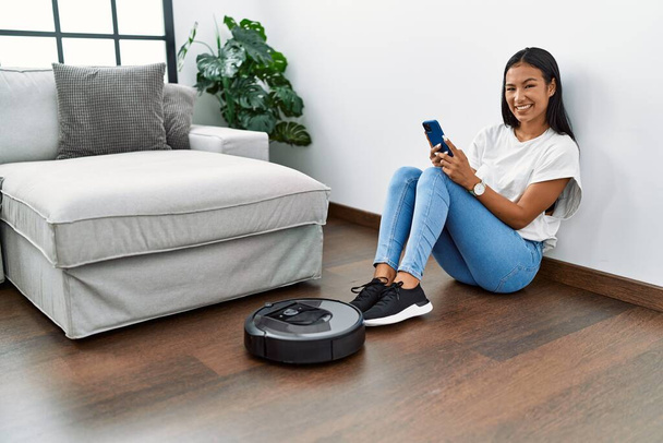 Young latin woman using smartphone cleaning using clean vacuum robot at home - Photo, Image