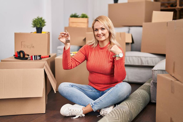 Blondynka trzymająca klucze nowego domu siedząca na podłodze wskazując palcem na siebie uśmiechniętą szczęśliwą i dumną  - Zdjęcie, obraz