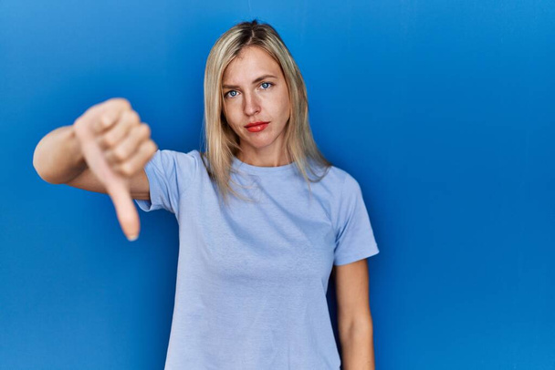 Beautiful blonde woman wearing casual t shirt over blue background looking unhappy and angry showing rejection and negative with thumbs down gesture. bad expression.  - Photo, Image