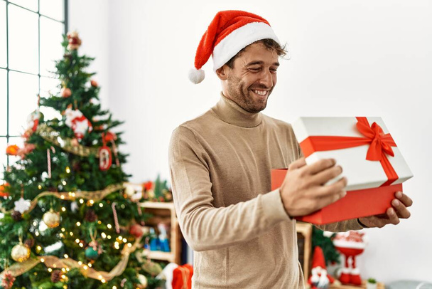 Joven hombre hispano sonriendo confiado sosteniendo regalo de Navidad en casa - Foto, Imagen