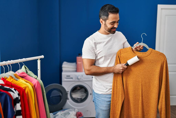 Young hispanic man cleaning sweater using hair pet roller at laundry room - Valokuva, kuva