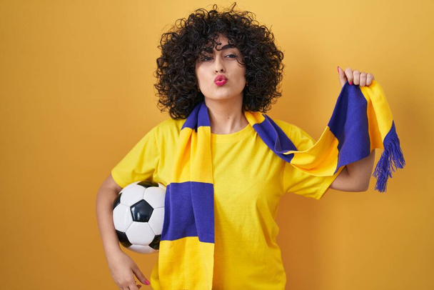 Mladá brunetka s kudrnatými vlasy fotbal chuligán drží míč při pohledu do kamery foukání polibek je krásná a sexy. výraz lásky.  - Fotografie, Obrázek