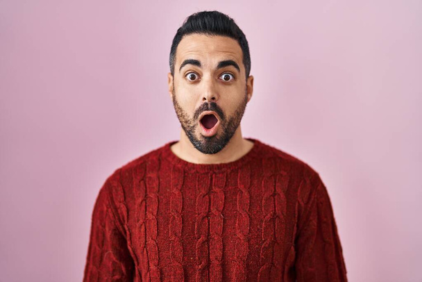 Молодий іспаномовний чоловік з бородою в повсякденному светрі над рожевим тлом боїться і шокований з несподіваним виразом, страхом і збудженим обличчям
.  - Фото, зображення