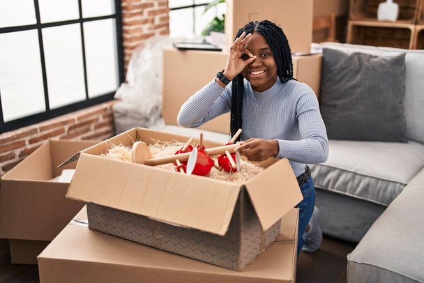 Αφροαμερικανή γυναίκα κινείται σε ένα νέο σπίτι ανοίγοντας κουτιά χαμογελώντας χαρούμενος κάνει εντάξει σημάδι με το χέρι στο μάτι κοιτάζοντας μέσα από τα δάχτυλα  - Φωτογραφία, εικόνα