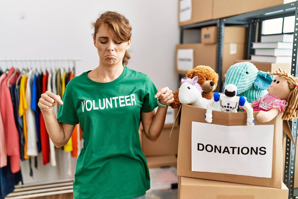 Красива біла жінка в волонтерській сорочці на пожертвуваннях стоїть, вказуючи на сум і засмученість, вказуючи напрямок пальцями, нещасний і пригнічений
.  - Фото, зображення