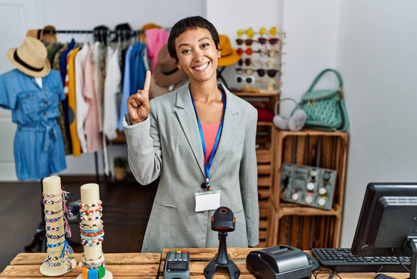 Jeune femme hispanique aux cheveux courts travaillant comme manager à la boutique de détail montrant et pointant vers le haut avec le doigt numéro un tout en souriant confiant et heureux.  - Photo, image