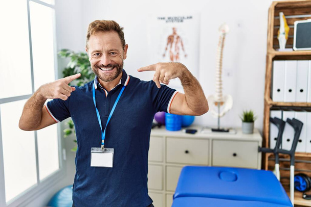 Middle age physiotherapist man working at pain recovery clinic smiling cheerful showing and pointing with fingers teeth and mouth. dental health concept.  - Photo, Image