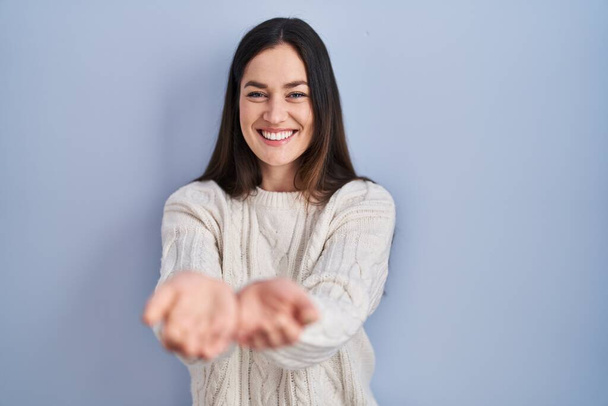 Nuori brunette nainen seisoo sininen tausta hymyillen kädet kämmenet yhdessä vastaanottaa tai antaa ele. hallussapito ja suojelu  - Valokuva, kuva