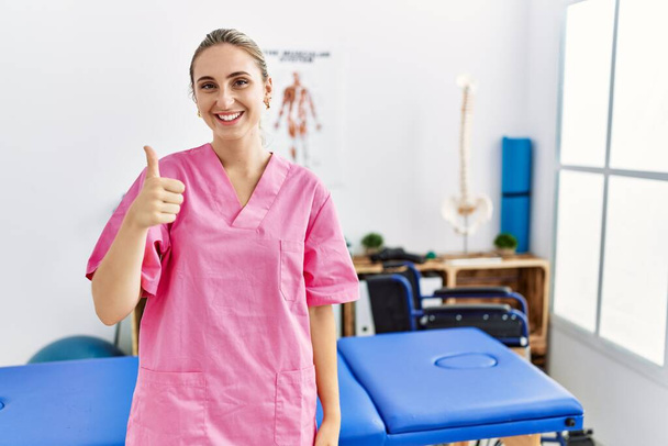 Young blonde woman working at pain recovery clinic doing happy thumbs up gesture with hand. approving expression looking at the camera showing success.  - Photo, Image