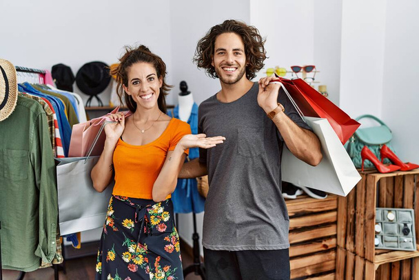Junges hispanisches Paar hält Einkaufstüten im Einzelhandel und lächelt fröhlich und zeigt mit der Handfläche in die Kamera.  - Foto, Bild