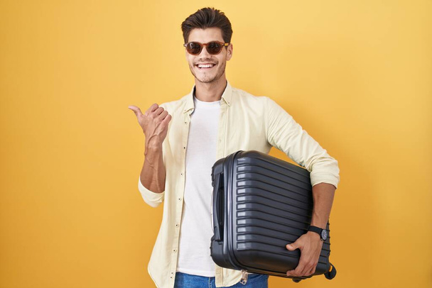Joven hombre hispano sosteniendo la maleta yendo de vacaciones de verano apuntando hacia atrás con la mano y los pulgares hacia arriba, sonriendo confiado  - Foto, Imagen
