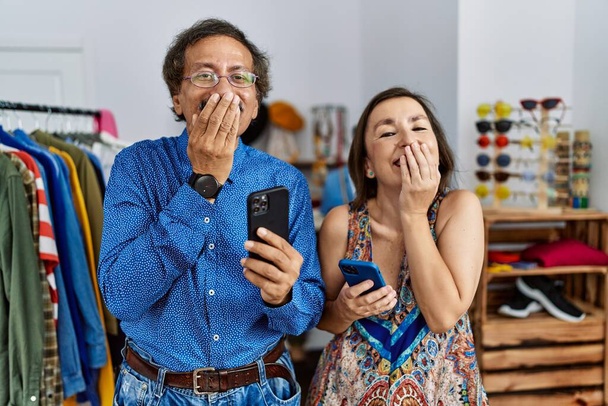 Middelbare leeftijd interraciale paar in de winkel met behulp van smartphone lachen en beschaamd giechelen bedekken mond met handen, roddel en schandaal concept  - Foto, afbeelding