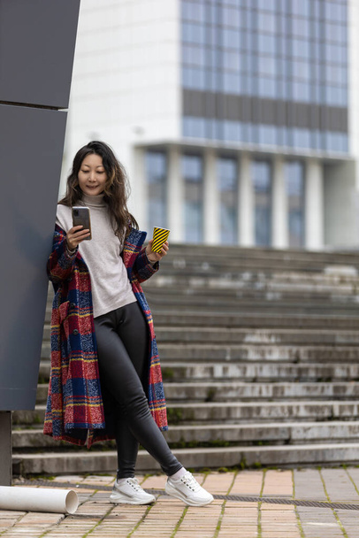 Attractive cheerful Asian woman in trench coat with coffee to walk happily with mobile phone outdoors. - Φωτογραφία, εικόνα