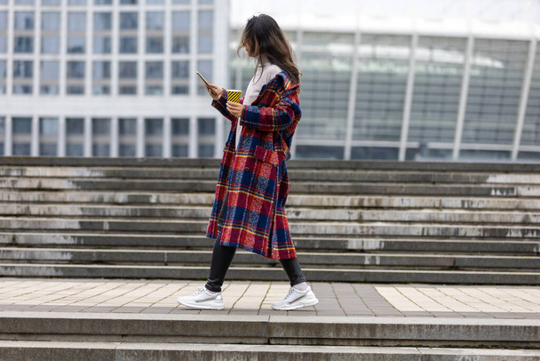 Mulher asiática alegre atraente em casaco de trincheira com café para andar feliz com telefone celular ao ar livre. - Foto, Imagem