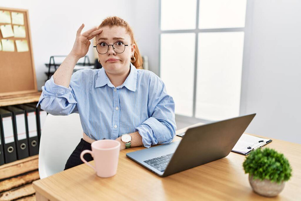 Giovane donna rossa che lavora in ufficio utilizzando il computer portatile preoccupato e stressato per un problema con la mano sulla fronte, nervoso e ansioso di crisi  - Foto, immagini