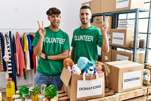 Ein junges schwules Paar im freiwilligen T-Shirt an einem Spendenstand lächelt mit glücklichem Gesicht in die Kamera und macht mit den Fingern ein Siegeszeichen. Nummer zwei.  - Foto, Bild