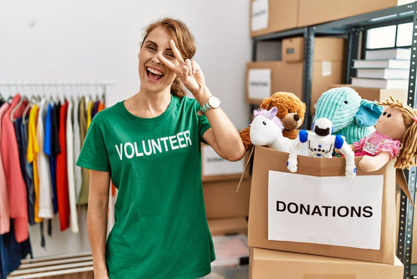 Hermosa mujer caucásica vistiendo camiseta voluntaria en el stand de donaciones haciendo símbolo de paz con los dedos sobre la cara, sonriendo alegre mostrando la victoria  - Foto, imagen