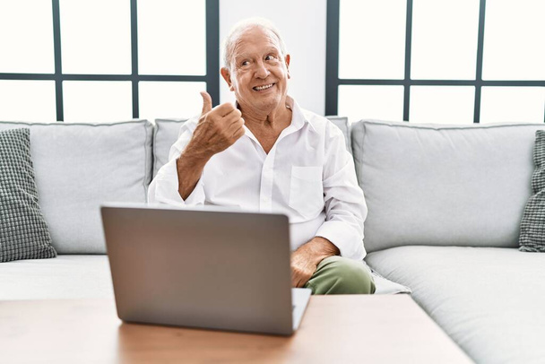 Старший использует ноутбук дома сидит на диване улыбается с счастливым лицом глядя и указывая на сторону с большим пальцем вверх.  - Фото, изображение