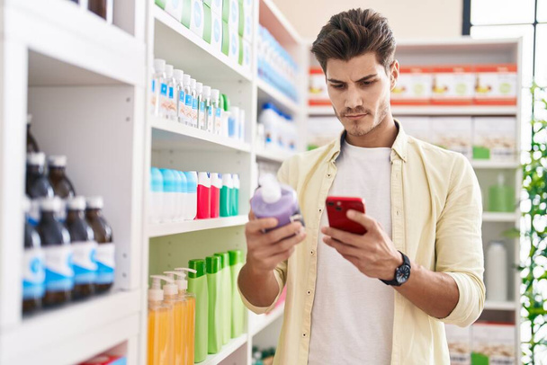 Молодой латиноамериканец клиент, использующий смартфон с гелевой бутылкой в аптеке - Фото, изображение