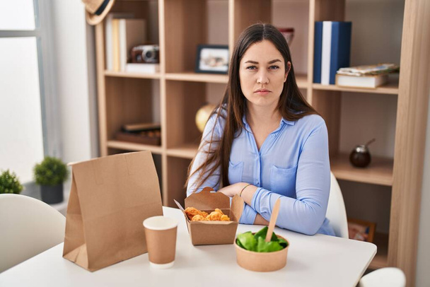 Junge brünette Frau, die zu Hause Essen zum Mitnehmen isst, denkt Haltung und nüchterner Ausdruck und sieht selbstbewusst aus  - Foto, Bild